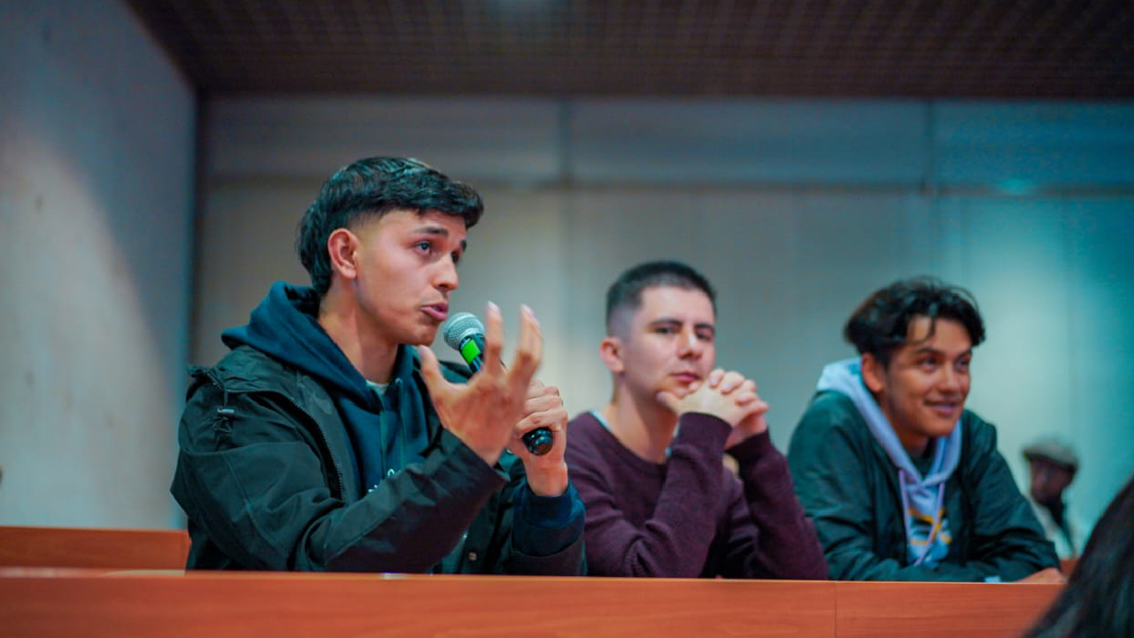 Joven participando activamente con micrófono en un espacio formativo del IDPAC, acompañado por otros asistentes en un auditorio.