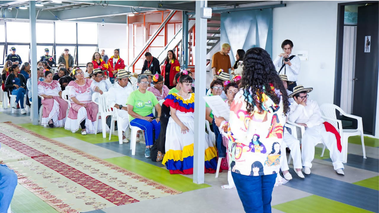 Personas reunidas en una jornada cultural en Bogotá, con asistentes vestidos con trajes tradicionales mientras escuchan a una mujer que lee en voz alta frente al grupo.