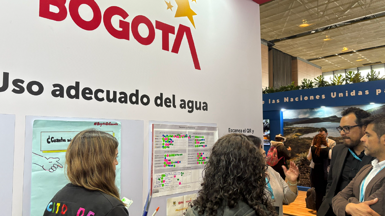 Funcionarias del IDPAC dialogan con asistentes sobre el uso adecuado del agua, frente a tableros participativos en la Cumbre Internacional de Sostenibilidad, destacando estrategias de educación ambiental y participación ciudadana.