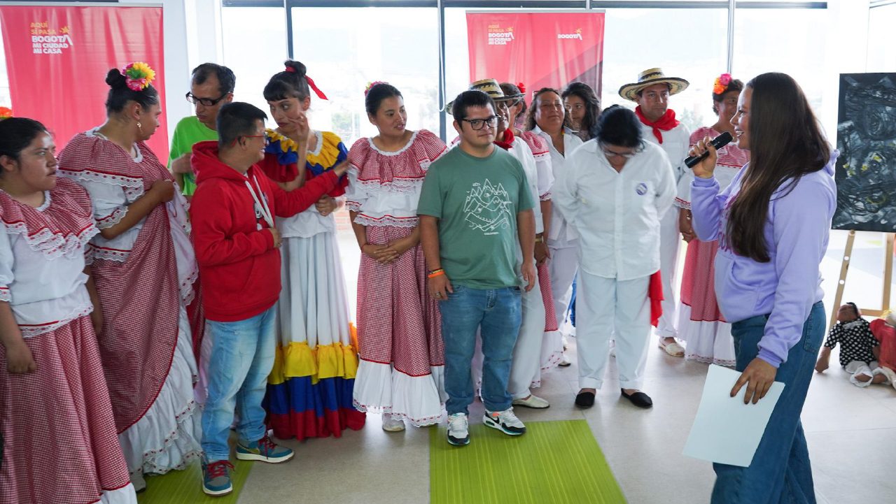 Grupo de personas en presentación cultural en Bogotá, con participantes en trajes tradicionales y una mujer dirigiendo la actividad con micrófono.