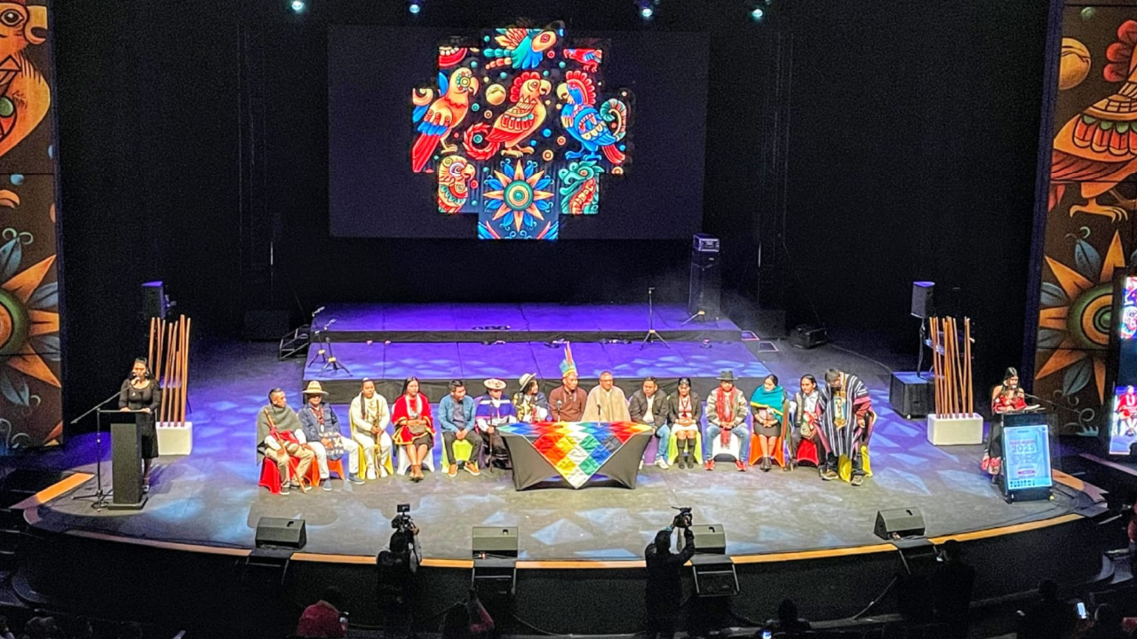 Representantes de distintos pueblos indígenas sentados en el escenario del Teatro Jorge Eliécer Gaitán, durante el Encuentro Distrital de Pueblos Indígenas 2025 en Bogotá. Al fondo se proyecta un símbolo colorido de identidad cultural