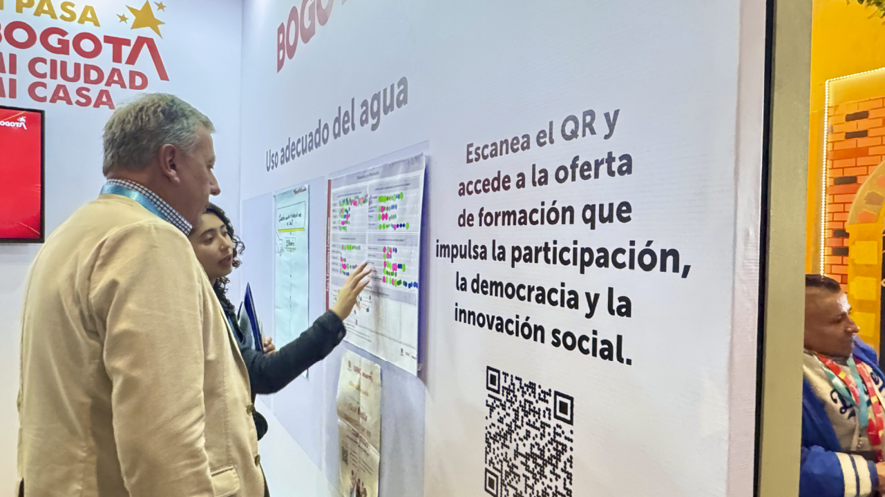 Persona explica a un visitante el ejercicio sobre el uso adecuado del agua en el stand del IDPAC durante la Cumbre de Sostenibilidad, destacando la oferta formativa en participación, democracia e innovación social.