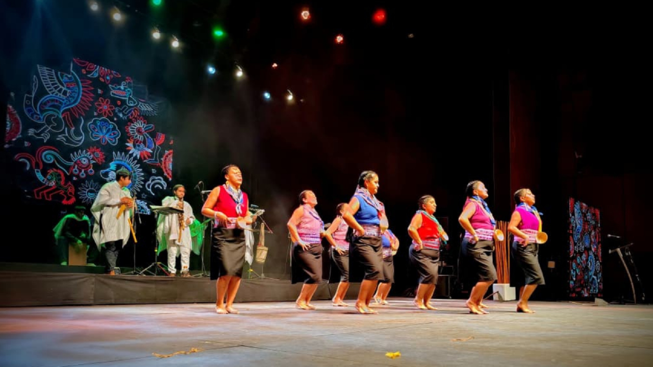Presentación artística de mujeres indígenas en el Encuentro Distrital de Pueblos Indígenas 2025. Ocho mujeres vestidas con atuendos tradicionales bailan en línea al ritmo de música ancestral interpretada en vivo, sobre un escenario iluminado y decorado con patrones culturales coloridos