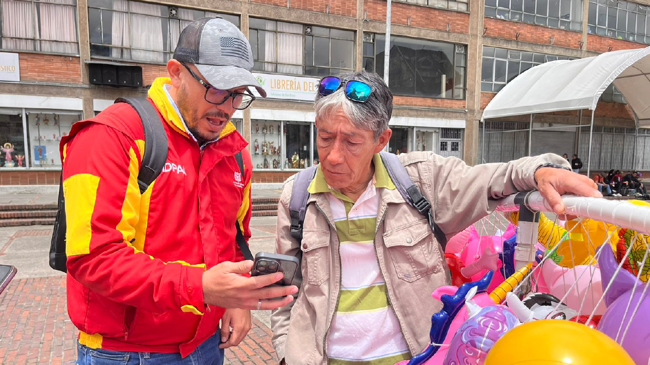 Servidor del IDPAC orienta a un vendedor informal en Bogotá mientras revisan información en un celular durante el proceso electoral de los Consejos Locales.