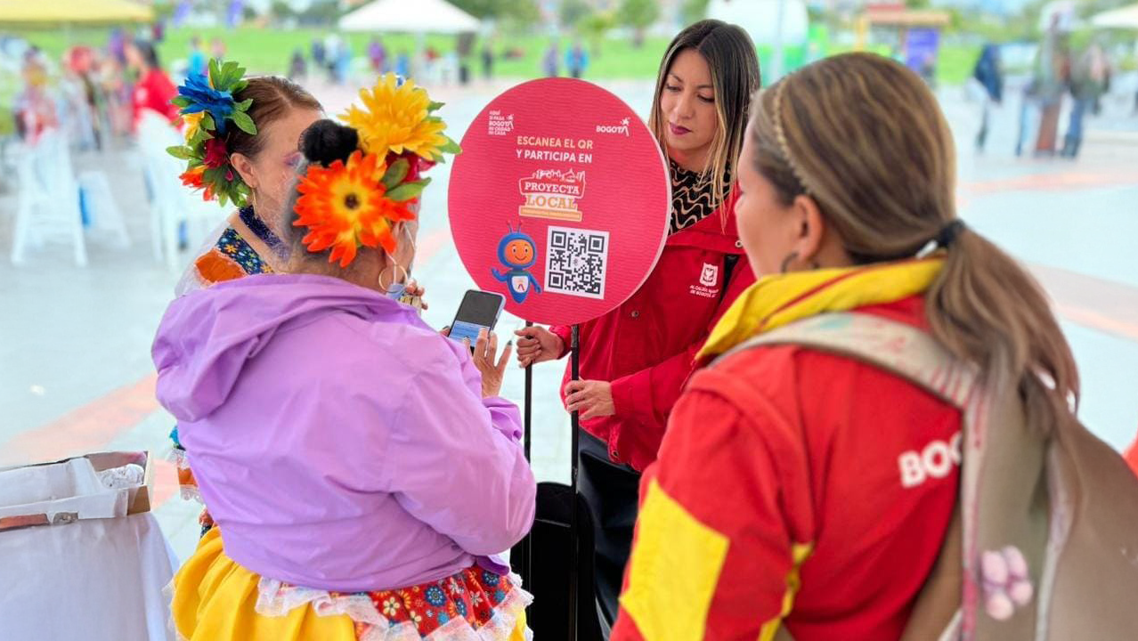 Gestoras del IDPAC orientan a una ciudadana durante una jornada de Proyecta Local, mostrando un cartel con código QR para participar en la priorización de iniciativas en Bogotá.