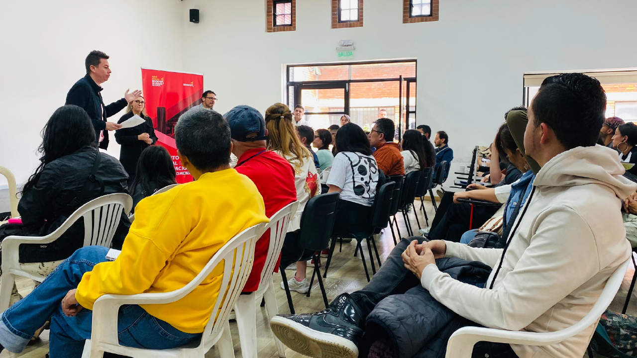 Personas participando en un encuentro comunitario del IDPAC dentro de un salón, escuchando a un facilitador al frente.