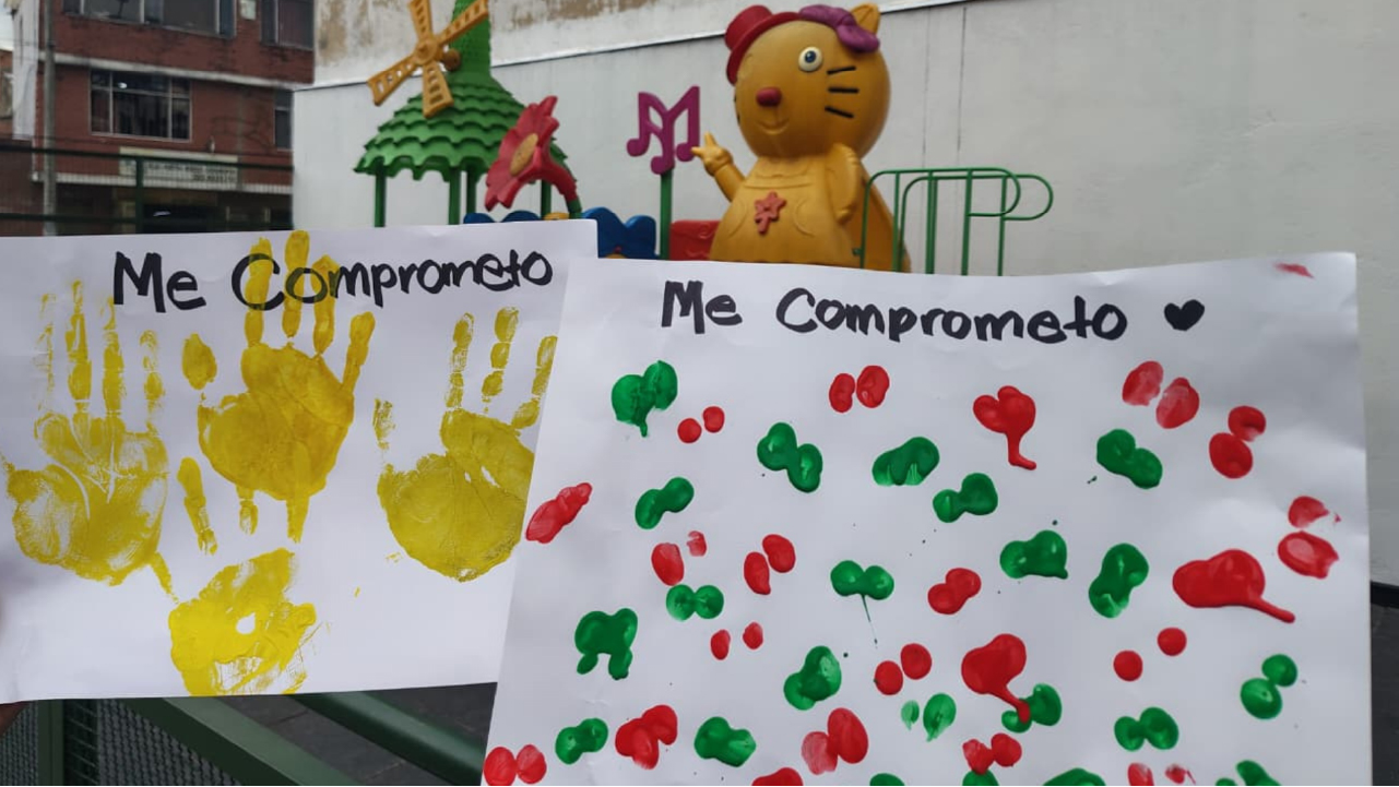 Dos carteleras con el texto "Me comprometo" decoradas con múltiples huellas de manos pintadas con témperas de diversos colores.