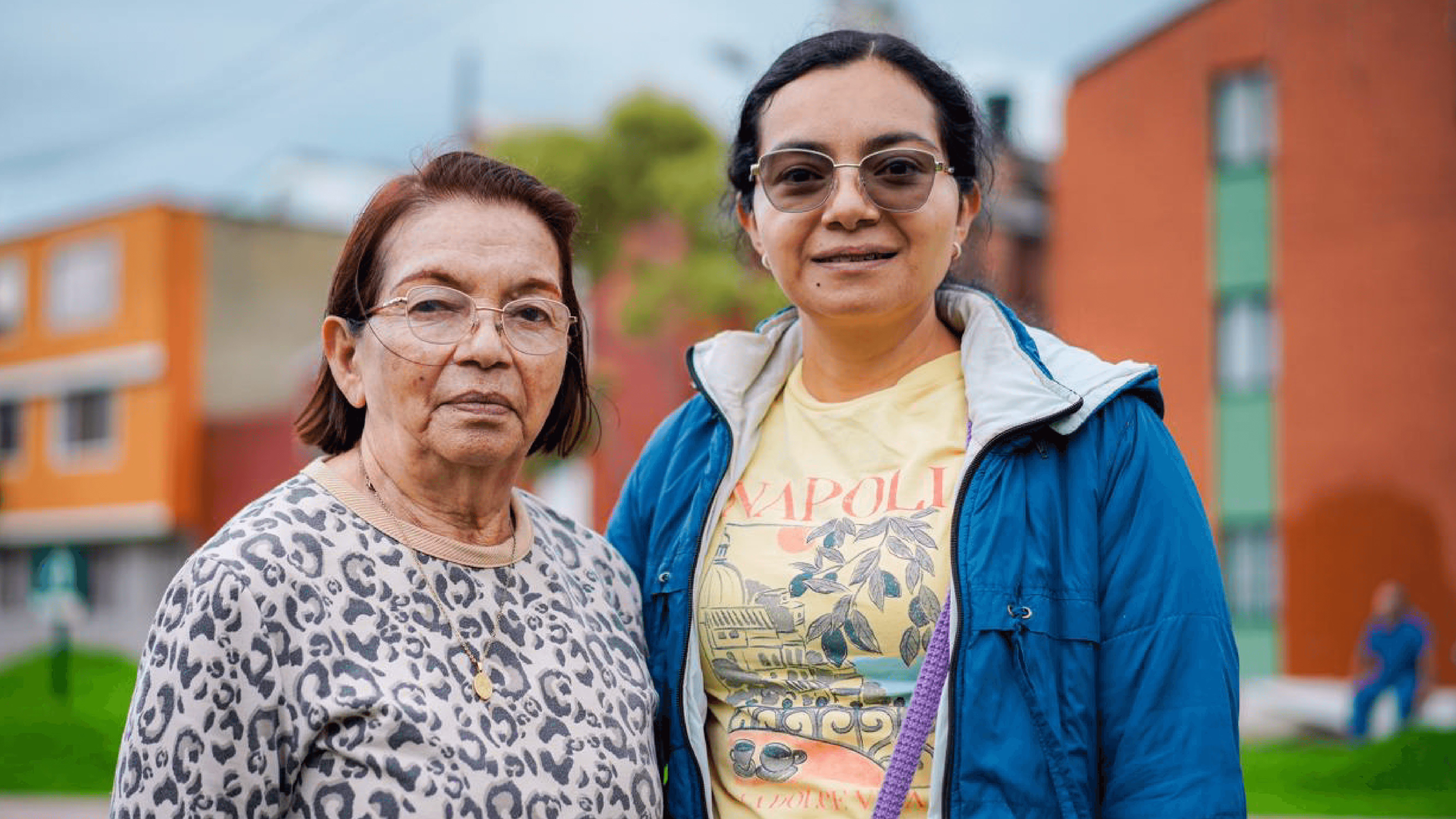Dos mujeres adultas posan juntas frente a un conjunto residencial en Bogotá, en un entorno urbano con edificios y zonas verdes