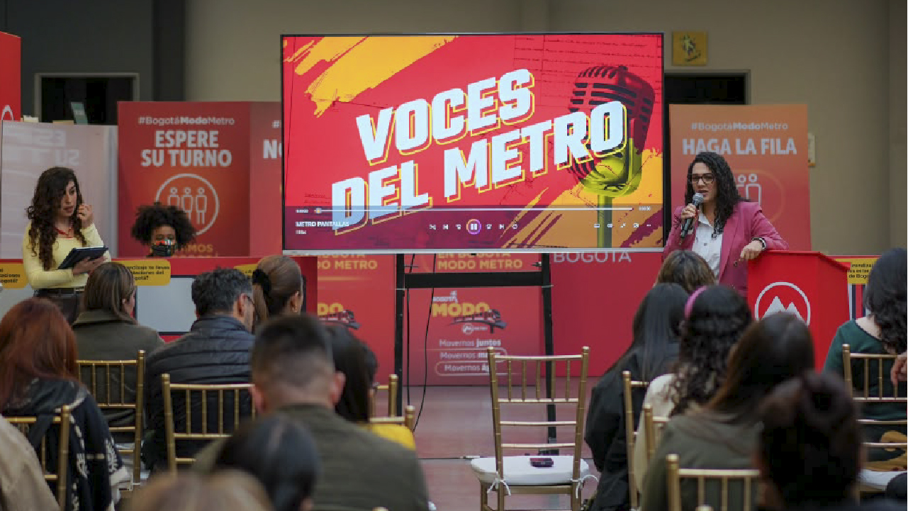 Presentadora habla al público durante el lanzamiento del pódcast 'Voces del Metro', proyectado en pantalla, en un evento de la estrategia Bogotá Modo Metro.