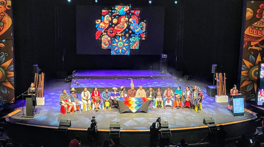 Representantes de distintos pueblos indígenas sentados en el escenario del Teatro Jorge Eliécer Gaitán, durante el Encuentro Distrital de Pueblos Indígenas 2025 en Bogotá. Al fondo se proyecta un símbolo colorido de identidad cultural