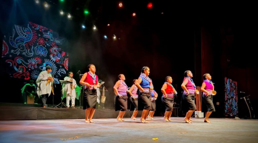 Presentación artística de mujeres indígenas en el Encuentro Distrital de Pueblos Indígenas 2025. Ocho mujeres vestidas con atuendos tradicionales bailan en línea al ritmo de música ancestral interpretada en vivo, sobre un escenario iluminado y decorado con patrones culturales coloridos