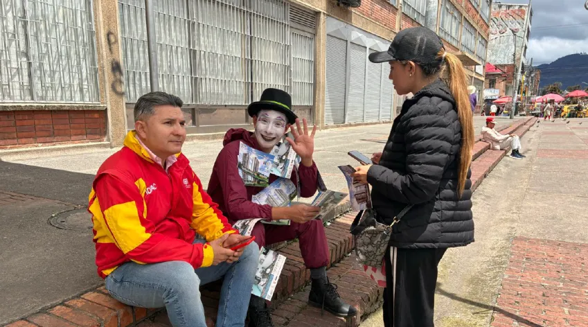 Gestores del IDPAC y un artista caracterizado como mimo entregan información a una ciudadana en vía pública durante jornada de participación en Bogotá.
