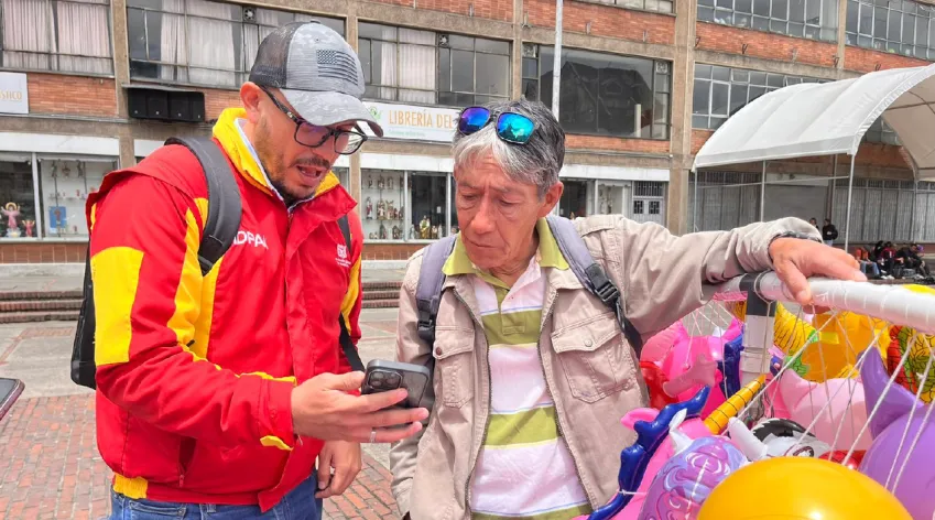 Servidor del IDPAC orienta a un vendedor informal en Bogotá mientras revisan información en un celular durante el proceso electoral de los Consejos Locales.