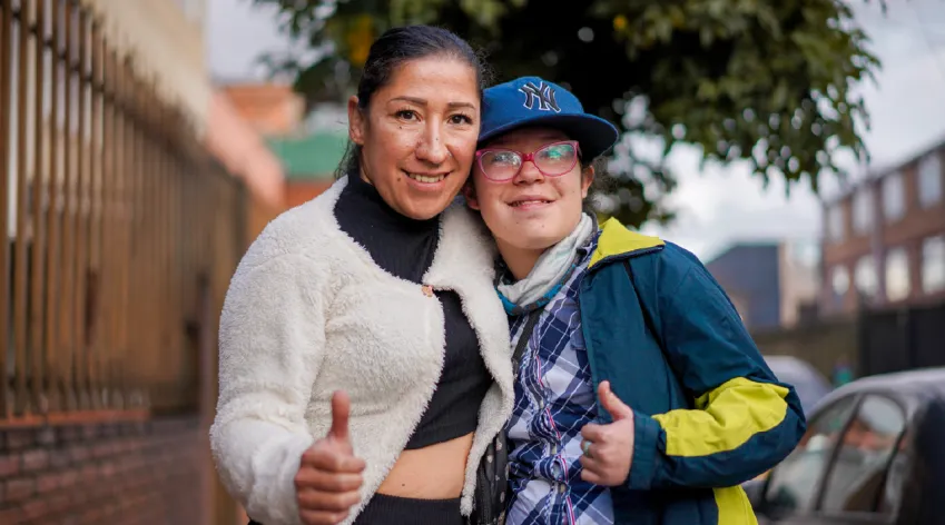 Dos personas sonríen y hacen pulgar arriba, abrazadas en una calle; retrato de inclusión y apoyo.