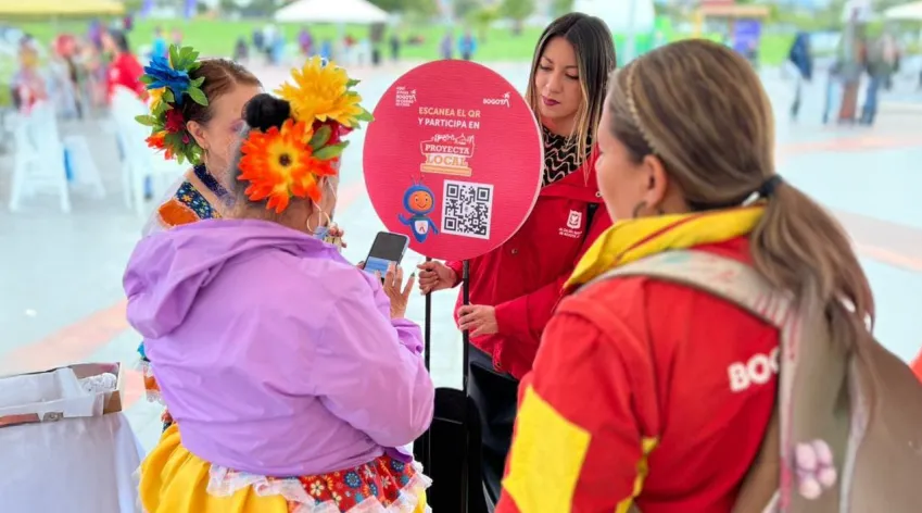 Gestoras del IDPAC orientan a una ciudadana durante una jornada de Proyecta Local, mostrando un cartel con código QR para participar en la priorización de iniciativas en Bogotá.