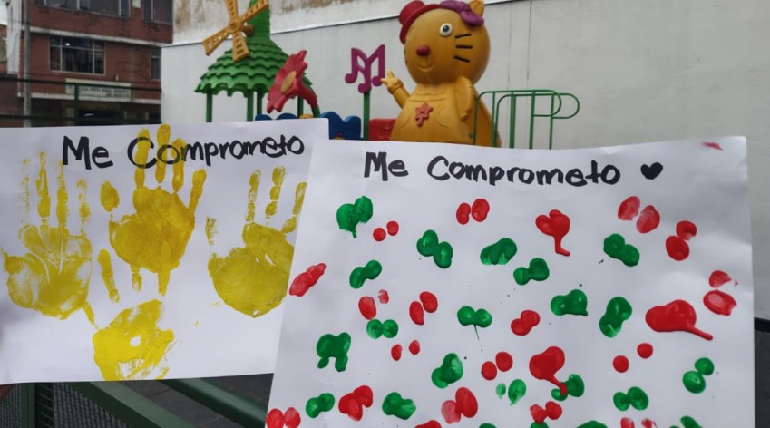 Dos carteleras con el texto "Me comprometo" decoradas con múltiples huellas de manos pintadas con témperas de diversos colores.