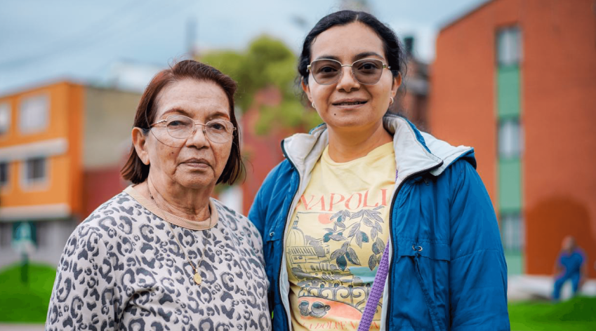 Dos mujeres adultas posan juntas frente a un conjunto residencial en Bogotá, en un entorno urbano con edificios y zonas verdes