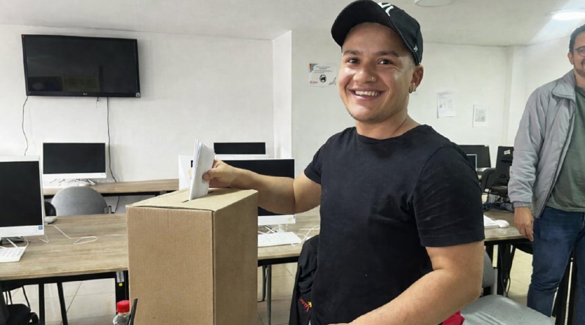 Imagen de un joven sonriente votando en una urna del IDPAC. En el fondo, las pantallas de computadora muestran la plataforma VOTEC y carteles con la marca institucional para la elección de propiedad horizontal en Bogotá hasta el 17 de abril.